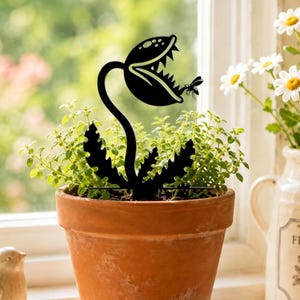 May include: A black metal plant stake featuring a silhouette of a carnivorous plant with a wide-open mouth and jagged teeth, capturing a small insect. The stake is inserted into a terracotta pot filled with green herbs, positioned near a window.