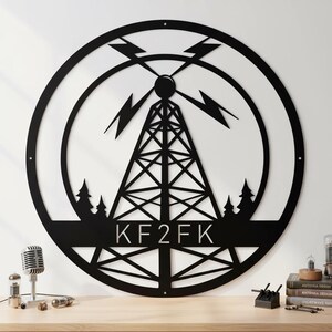 Peut inclure: Décoration murale en métal noir représentant une tour radio dans un cercle, avec des éclairs et des arbres. Les lettres "KF2FK" sont affichées en bas. L'art est affiché sur un mur blanc au-dessus d'une surface en bois.