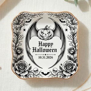 May include: A square ceramic trinket dish with a gold-toned rim featuring a black and white illustration of a winged pumpkin, roses, skulls, and bare trees. The center text reads Happy Halloween and 10.31.2026 in a decorative font style.