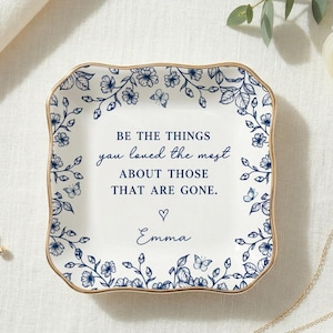 May include: A square white ceramic trinket dish with scalloped edges and a gold-toned rim. The dish features a blue floral and butterfly border pattern and the text: BE THE THINGS you loved the most ABOUT THOSE THAT ARE GONE. A heart and Emma.