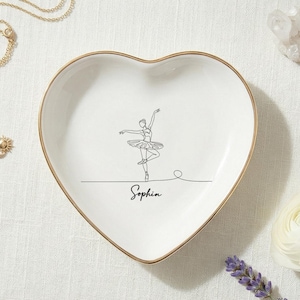 May include: A white heart-shaped ceramic trinket dish with a gold-colored rim. The center features a black line art illustration of a ballerina in a dance pose, with the name Sophin written in a cursive script font directly below the figure.