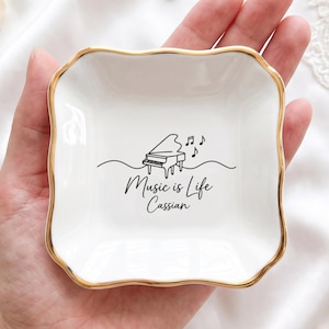 May include: A small, square white ceramic trinket dish with a scalloped edge and a gold-painted rim. The center features a black line drawing of a grand piano with musical notes and the text "Music is Life" followed by the name "Cassian".