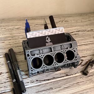 May include: A gray, 3D-printed engine block desk organizer holding pens, business cards, and other office supplies. The business card has the text "3DSYS" on it. The organizer sits on a wooden surface.