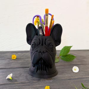 Puede incluir: Un organizador de escritorio negro con forma de bulldog que contiene útiles de arte. El organizador contiene pinceles, tijeras y bolígrafos. Flores amarillas y pétalos blancos están esparcidos sobre una superficie de madera oscura.