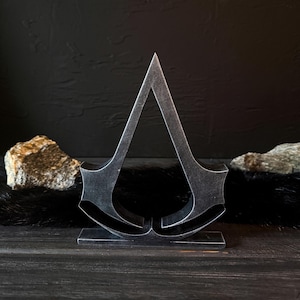 May include: A metallic silver sculpture of a stylized, upward-pointing triangle with curved elements at the base. The sculpture is set on a rectangular base and placed on a dark wooden surface. Two rocks are on either side.