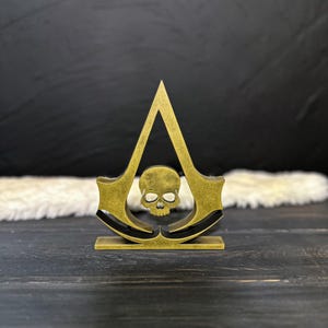 Può includere: Un emblema stilizzato color oro con un teschio all'interno di una forma triangolare, con elementi curvi su entrambi i lati. Il design è posto su una base rettangolare e lo sfondo è scuro.