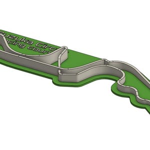 Puede incluir: Modelo impreso en 3D, verde y gris, del circuito de carreras de Suzuka. El trazado de la pista es gris sobre una base verde. El texto "Suzuka Circuit Racing Course" es visible.