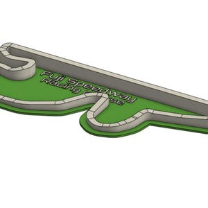 Puede incluir: Un cortador de galletas verde y gris con la forma del circuito de carreras Fuji Speedway. El texto "Fuji Speedway Racing Course" está impreso en la superficie verde. La pista está delineada en gris.