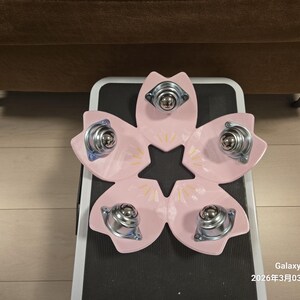 Könnte beinhalten: Ein rosa, blütenförmiges Objekt mit fünf Blütenblättern, die jeweils mit einer silbernen Lenkrolle ausgestattet sind. Die Blütenblätter haben gelbe Akzente. Das Objekt befindet sich auf einer schwarz-weißen Oberfläche.