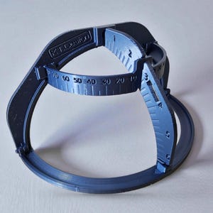 Può includere: Uno strumento di misurazione in plastica blu con un design curvo. Presenta una scala circolare con numeri da 10 a 70 e il testo "K.T. Design". Lo strumento ha bracci curvi e una base.