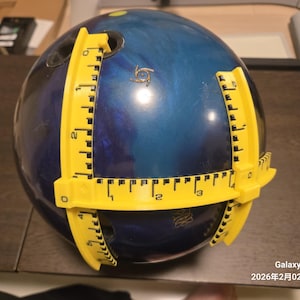 以下が含まれることがあります： 青と黒のボウリングボールと黄色の測定ツール。 測定ツールはボールの周りに配置され、サイズを測定します。 ボールには3つの指穴があります。 テキスト「Galaxy Z Fold6 2026年2月02日 22:38」が表示されます。