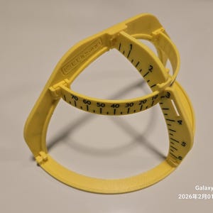 Puede incluir: Una herramienta de medición de plástico amarillo con brazos curvos y una base circular. La herramienta tiene marcas de medición impresas en negro, que van de 0 a 70. La herramienta está diseñada para medir y marcar. El texto "K.L. Design" es visible.