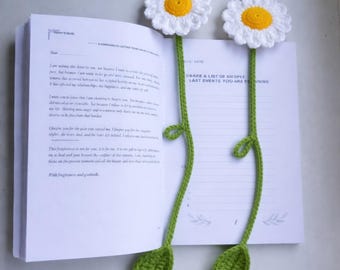 Könnte beinhalten: Zwei gehäkelte Gänseblümchen-Lesezeichen mit weißen Blütenblättern und gelben Zentren. Die grünen Stiele haben Blätter und sind in einem offenen Buch platziert. Die Seiten des Buches sind sichtbar, mit Text und Linien.