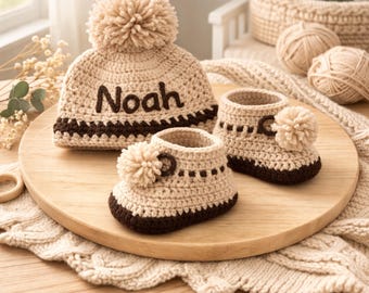 Könnte beinhalten: Gehäkeltes Baby-Set in Beige und Braun, bestehend aus einer Mütze mit Bommel und dem Namen "Noah" sowie zwei Babyschuhen, ebenfalls mit Bommeln. Das Set wird auf einer Holzoberfläche präsentiert.