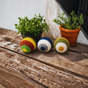K&ouml;nnte beinhalten: Drei kugelf&ouml;rmige H&auml;kelb&auml;lle mit unterschiedlichen Farbmustern. Der erste Ball hat konzentrische Ringe in Gelb, Gr&uuml;n und Kastanienbraun. Der zweite Ball zeigt Ringe in Blau, Grau und Wei&szlig;. Der dritte Ball ist in Gelb, Wei&szlig; und Olivgr&uuml;n gehalten.