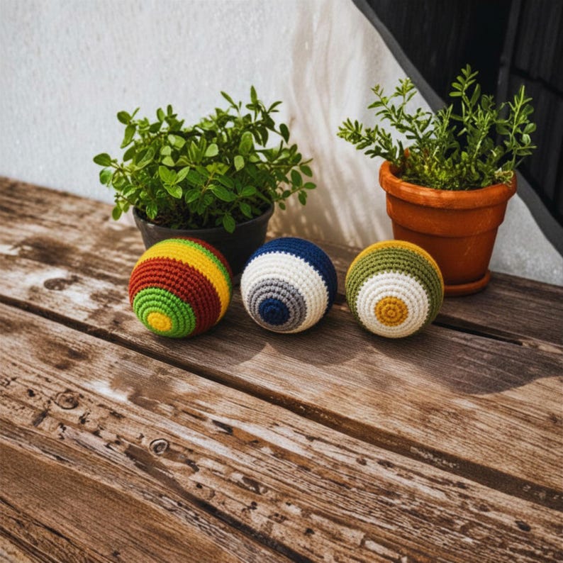 K&ouml;nnte beinhalten: Drei kugelf&ouml;rmige H&auml;kelb&auml;lle mit unterschiedlichen Farbmustern. Der erste Ball hat konzentrische Ringe in Gelb, Gr&uuml;n und Kastanienbraun. Der zweite Ball zeigt Ringe in Blau, Grau und Wei&szlig;. Der dritte Ball ist in Gelb, Wei&szlig; und Olivgr&uuml;n gehalten.
