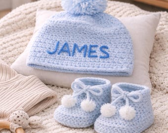 Könnte beinhalten: Hellblaues Baby-Set aus Häkelarbeit, bestehend aus einer Mütze mit dem Namen "JAMES" in Blau gestickt, einem passenden Bommel und einem Paar Babyschuhe mit weißen Bommeln und Schleifen. Die Artikel liegen auf einer cremefarbenen Oberfläche.