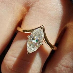 May include: A gold ring with a marquise-cut diamond. The diamond is set in a V-shaped design, and the band is a simple, polished gold. The ring is shown on a finger, with a dark background.