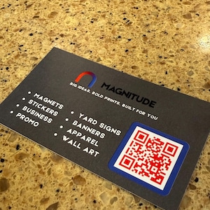 Könnte beinhalten: Eine Visitenkarte mit dem Wort "MAGNITUDE" und einem QR-Code. Die Karte listet Dienstleistungen wie Magnete, Aufkleber, Business, Promo, Werbeschilder, Banner, Bekleidung und Wandkunst auf. Die Karte liegt auf einer gesprenkelten braunen Oberfläche.