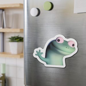 Könnte beinhalten: Ein grüner Froschmagnet mit weißem Rand ist an einem Kühlschrank aus Edelstahl befestigt. Der Frosch hat rosa Augen und winkt. Zwei runde Magnete, einer weiß und einer grün, befinden sich ebenfalls auf dem Kühlschrank.