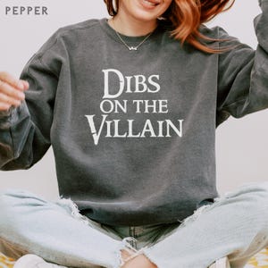 K&ouml;nnte beinhalten: Ein dunkelgraues Sweatshirt mit dem wei&szlig;en Text "Dibs on the Villain". Das Sweatshirt hat einen Rundhalsausschnitt und lange &Auml;rmel. Das Wort "Pepper" ist oben links zu sehen.