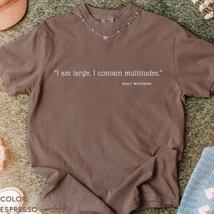 May include: Espresso-colored t-shirt featuring the quote "I am large. I contain multitudes." by Walt Whitman. The shirt has a crew neck and a delicate butterfly necklace. The fabric is soft and slightly wrinkled.