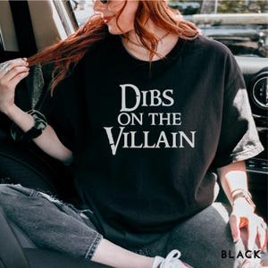 K&ouml;nnte beinhalten: Schwarzes T-Shirt mit dem wei&szlig;en Text "Dibs on the Villain". Das Wort "BLACK" ist unten rechts im Bild aufgedruckt. Das T-Shirt wird von einer Person getragen.