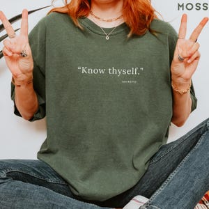 May include: Moss green sweatshirt with the quote "Know thyself." in white text. The quote is attributed to Socrates. The sweatshirt is a relaxed fit and is paired with blue jeans.