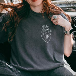 May include: Pepper-colored t-shirt featuring a white anatomical heart design on the chest. The shirt has a crew neck and short sleeves. The color is listed as "Pepper".