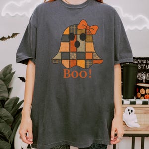 Könnte beinhalten: Dunkelgraues T-Shirt mit einem Patchwork-Geisterdesign, einer orangefarbenen Schleife und dem Wort "Boo!" in Orange. Der Geist hat ein Patchwork-Design in Orange-, Braun- und Beigetönen. Ein lustiges Halloween-T-Shirt.
