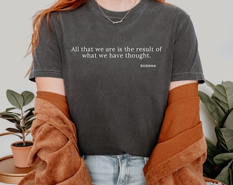 Può includere: T-shirt grigio scuro con la citazione "Tutto ciò che siamo è il risultato di ciò che abbiamo pensato" di Buddha. La maglietta è indossata con una giacca color ruggine e jeans blu. La parola "PEPPER" è in alto a destra.