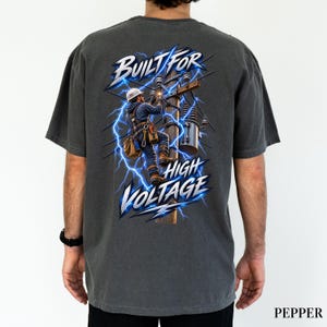 May include: A charcoal gray t-shirt featuring a graphic on the back of a utility worker in a hard hat climbing a wooden utility pole. The design includes blue lightning bolts and the text BUILT FOR HIGH VOLTAGE in a bold, stylized font.