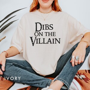K&ouml;nnte beinhalten: Elfenbeinfarbenes T-Shirt mit dem schwarzen Text "Dibs on the Villain". Das Shirt hat eine lockere Passform und wird mit einer blauen Jeans kombiniert. Das Wort "IVORY" ist unten links auf dem Shirt aufgedruckt.