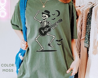 Peut inclure: T-shirt vert mousse avec un motif de squelette noir jouant de la guitare, portant un chapeau haut de forme, avec un croissant de lune et des chauves-souris. Le texte "COLOR: MOSS" est visible.