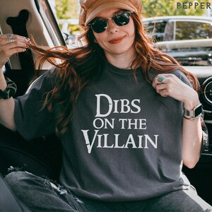 K&ouml;nnte beinhalten: Dunkelgraues T-Shirt mit dem wei&szlig;en Text "Dibs on the Villain". Das Shirt hat einen Rundhalsausschnitt und kurze &Auml;rmel. Die Person auf dem Bild tr&auml;gt eine Sonnenbrille und einen Hut.