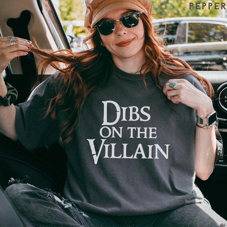 K&ouml;nnte beinhalten: Dunkelgraues T-Shirt mit dem wei&szlig;en Text "Dibs on the Villain". Das Shirt hat einen Rundhalsausschnitt und kurze &Auml;rmel. Die Person auf dem Bild tr&auml;gt eine Sonnenbrille und einen Hut.