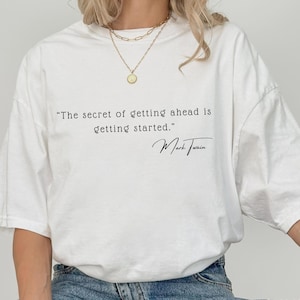 May include: White t-shirt with the quote "The secret of getting ahead is getting started." attributed to Mark Twain in black script. The shirt is a casual style, suitable for everyday wear.