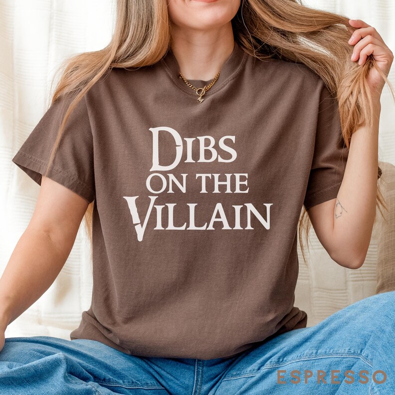 K&ouml;nnte beinhalten: Espressofarbenes T-Shirt mit dem wei&szlig;en Text "Dibs on the Villain". Das Shirt hat einen Rundhalsausschnitt und kurze &Auml;rmel. Das Model tr&auml;gt eine blaue Jeans.