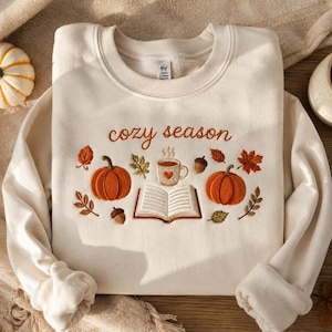 May include: A cream-colored crewneck sweatshirt featuring embroidered autumn-themed designs including two orange pumpkins, an open book, a mug with a heart, and scattered leaves and acorns. The text "cozy season" is embroidered in brown script.
