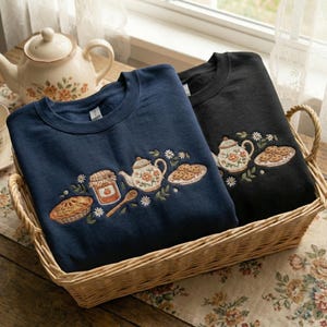 May include: Two crewneck sweatshirts, navy and black, sit in a wicker basket. Each has an embroidered design of a teapot, biscuits, pie, jam jar, and spoon. The sweatshirts are part of a tea-themed collection, perfect for a cosy aesthetic.