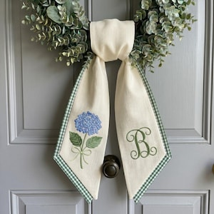 May include: Cream-colored door swag with a blue hydrangea embroidery and a green gingham trim. The swag is adorned with a green monogram letter "B". The swag is hanging on a gray door with a wreath.
