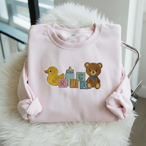 May include: A light pink sweatshirt featuring embroidered baby-themed graphics. The design includes a yellow duck, a baby bottle, a brown teddy bear with a blue bow, and blocks spelling "NICU". The sweatshirt is displayed on a white furry surface.