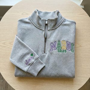 May include: A gray quarter-zip sweatshirt with the embroidered text "MARDI GRAS" in purple, yellow, and green. The sleeve features the name "olivia" embroidered in purple with a green heart. The sweatshirt is folded on a light wood surface.