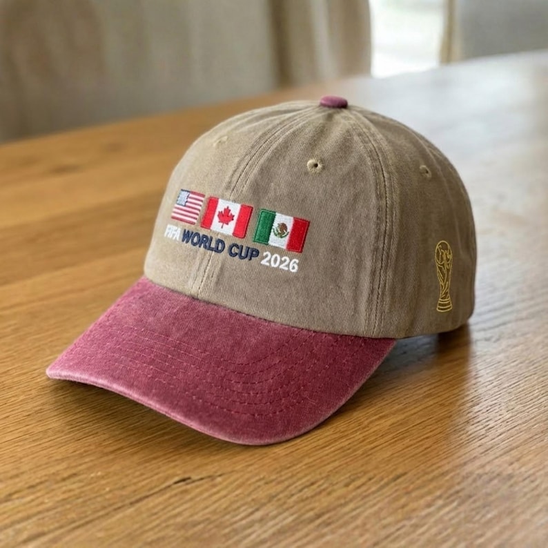 May include: A tan baseball cap with a maroon bill and top button. The cap features embroidered flags of the USA, Canada, and Mexico, with the text "FIFA WORLD CUP 2026." A gold FIFA trophy is embroidered on the side.