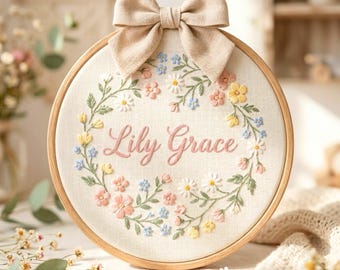 May include: Embroidered hoop art featuring the name "Lily Grace" in pink script, surrounded by a floral wreath of pink, yellow, blue, and white flowers. A beige bow is attached at the top of the wooden hoop.