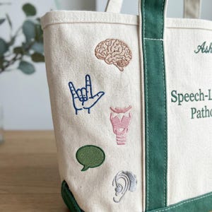 Puede incluir: Bolso tote de lona color crema con ribetes y asas verdes. La bolsa presenta parches bordados de un cerebro, un gesto con la mano, un bocadillo, una laringe y una oreja. El texto en la bolsa dice "Speech-Language Pathology".