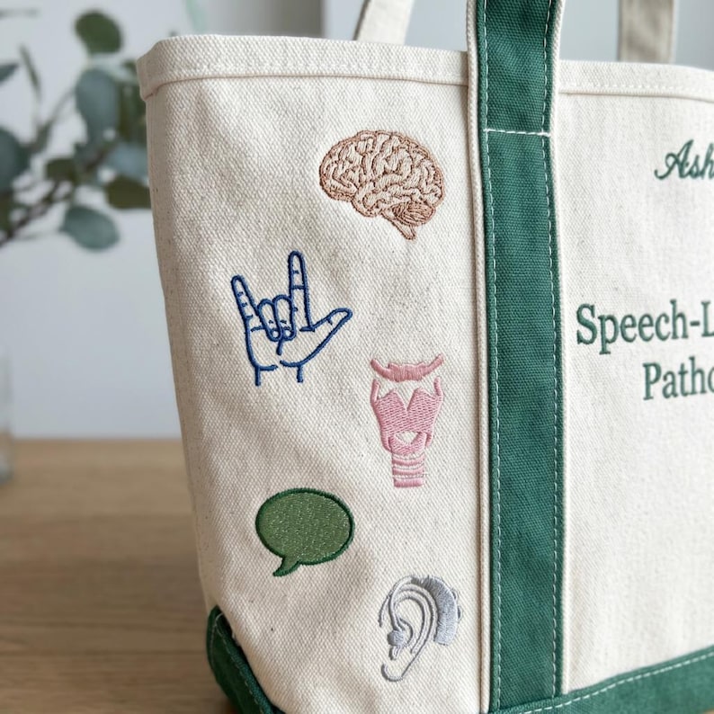 Puede incluir: Bolso tote de lona color crema con ribetes y asas verdes. La bolsa presenta parches bordados de un cerebro, un gesto con la mano, un bocadillo, una laringe y una oreja. El texto en la bolsa dice "Speech-Language Pathology".