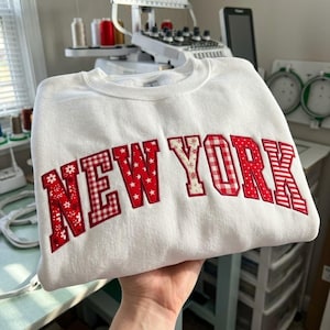 May include: A white crewneck sweatshirt featuring the text NEW YORK in large, red, fabric-appliqued letters. Each letter displays a different pattern, including floral, gingham, star, and striped designs, stitched onto the front of the garment.