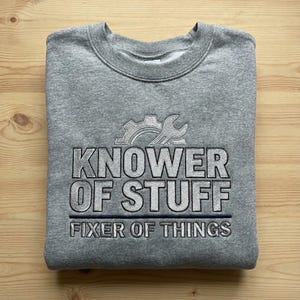 May include: A gray sweatshirt with the text "KNOWER OF STUFF FIXER OF THINGS" in a bold, outlined font. Above the text is a gear and wrench design. The sweatshirt is folded neatly on a wooden surface.