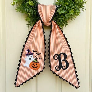 May include: A decorative door hanger featuring a green leafy wreath base with two long, orange and white striped fabric tails. One tail displays an embroidered white ghost and orange pumpkin, while the other features a large black letter B.